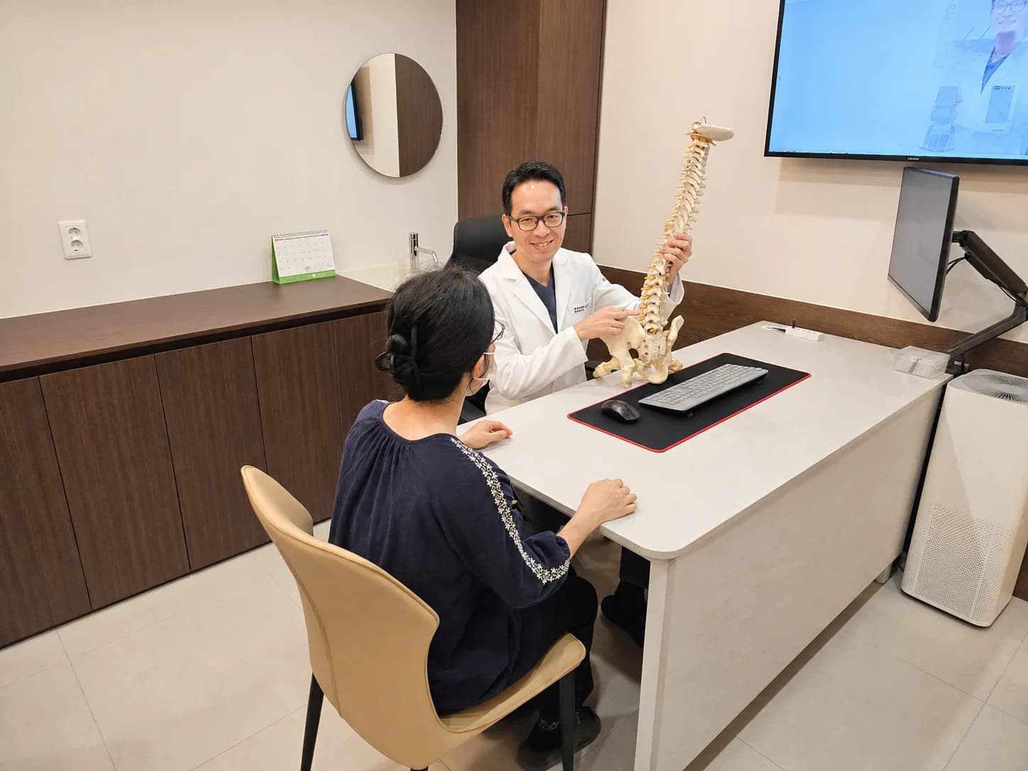 대표원장이 척추 모형으로 환자에게 추나 치료 과정을 설명하는 진료 상담 장면